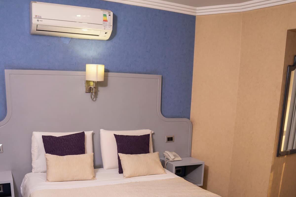 A hotel room with a large bed, an air conditioning unit, a bedside lamp, a nightstand, and a telephone.