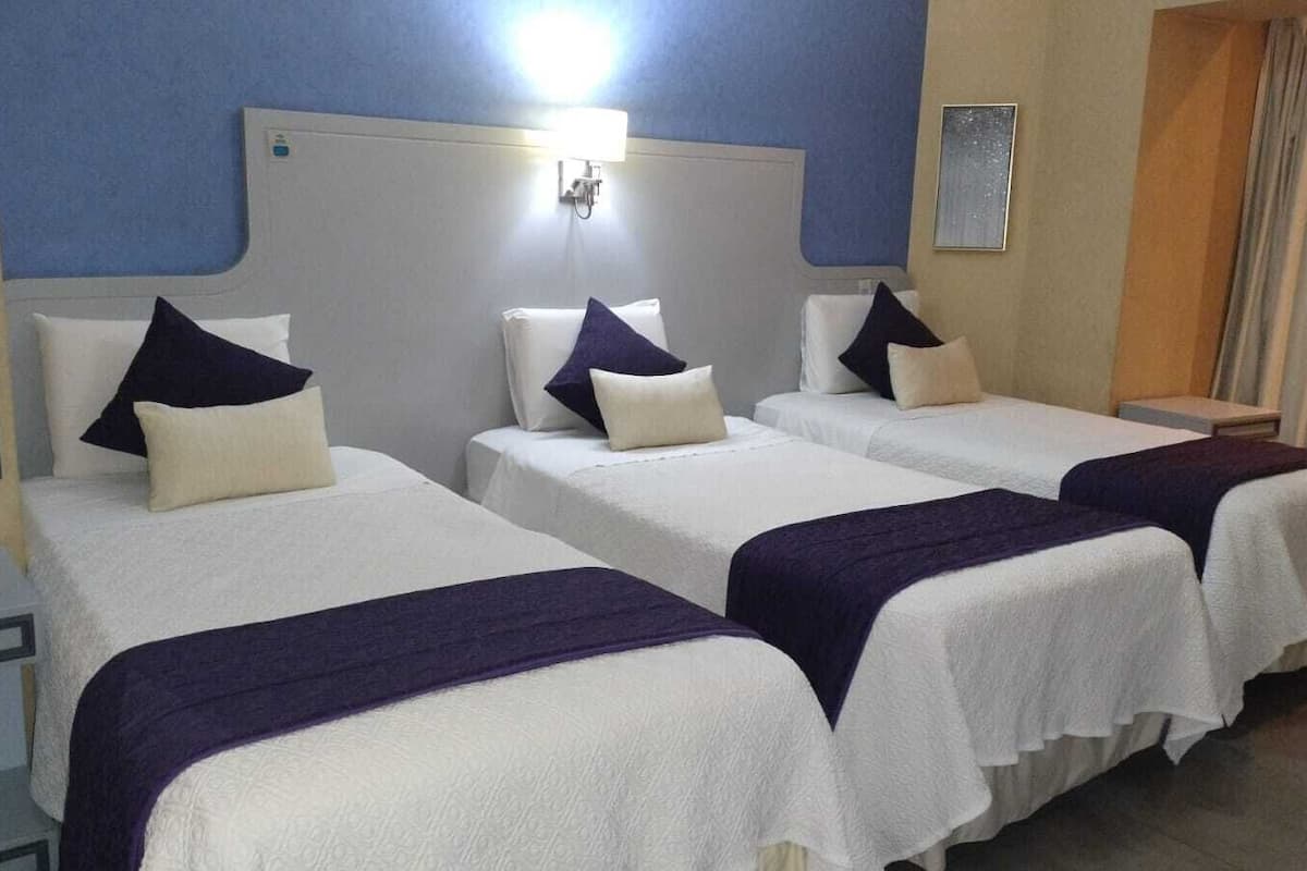 A hotel room with three beds, each with a white bedspread and a dark-colored throw blanket. There is a bedside table on the left, a wall-mounted lamp above the beds, and a small framed picture on the wall.