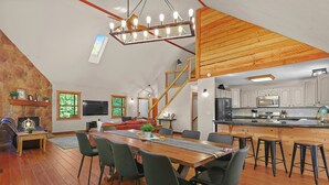 A spacious loft with a dining area, kitchen, and living space featuring wooden beams, a stone wall, and a large window.