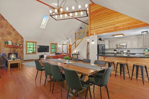 A spacious loft with a dining area, kitchen, and living space featuring wooden beams, a stone wall, and a large window.