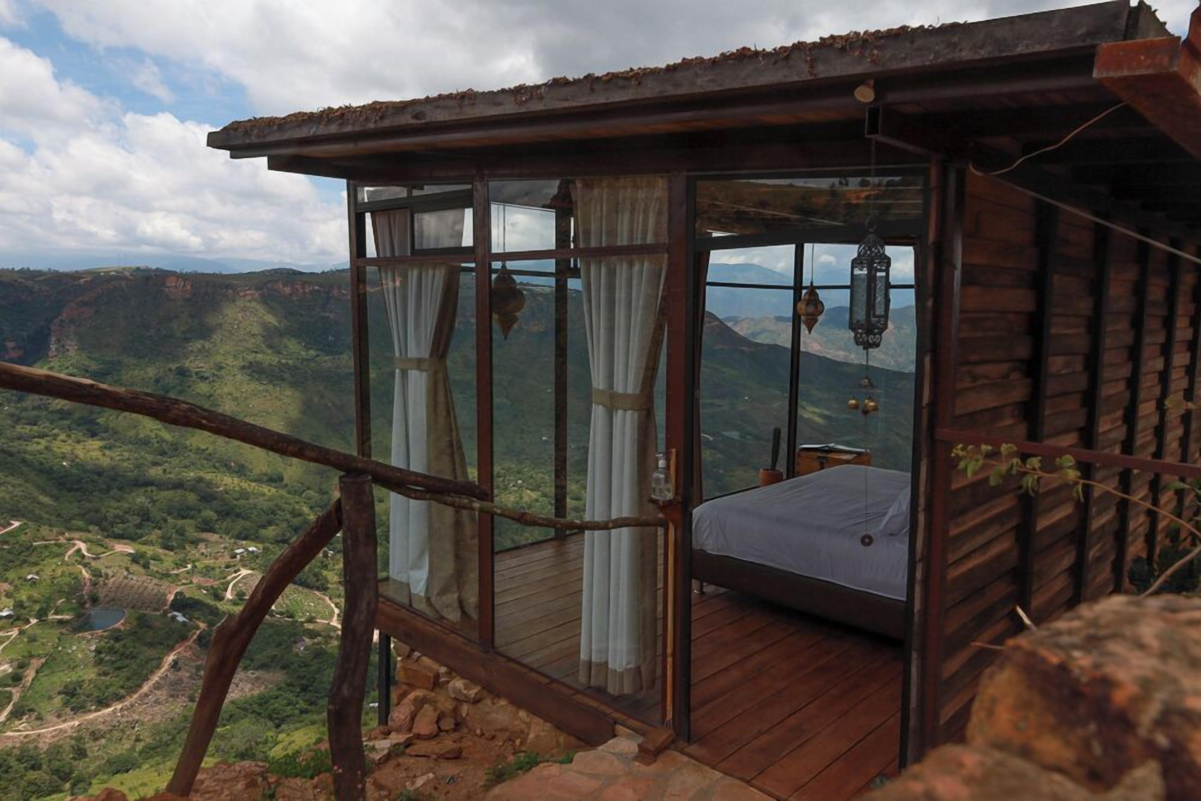 A wooden cabin with a bed, large windows, and a view of a mountainous landscape.