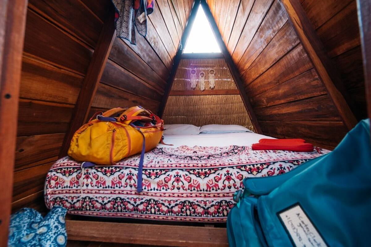 A small, triangular attic room with wooden walls and a sloped ceiling. There is a bed with a patterned bedspread and pillows, a yellow bag, and a red item on the bed.
