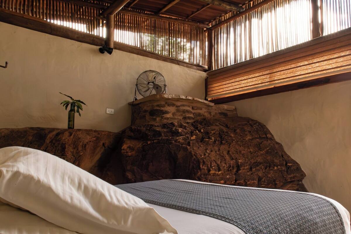 A room with a bed, a fan, and a stone wall.