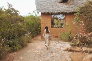 A woman walking on a stone pathway towards a thatched-roof building surrounded by lush greenery.