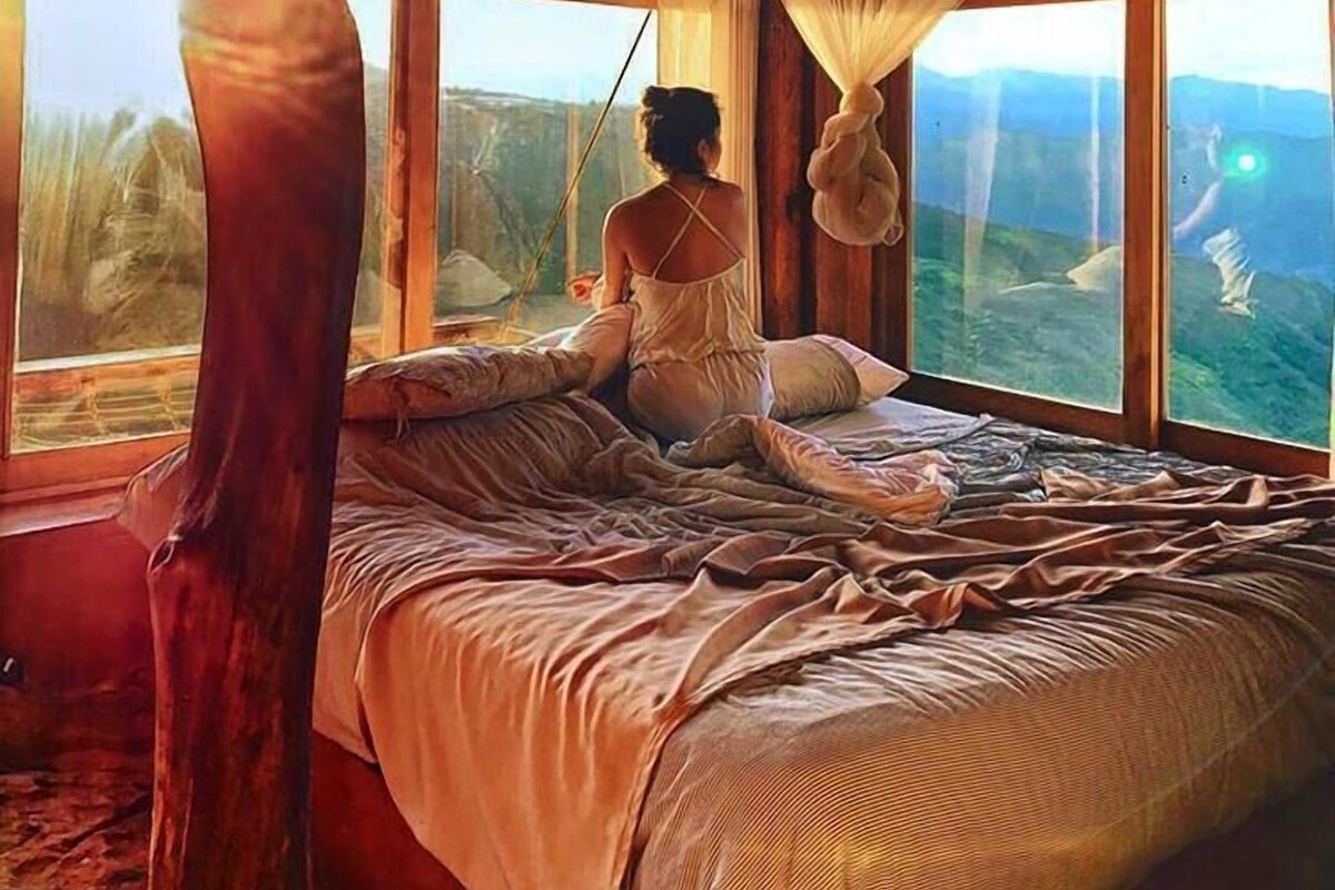 A room with a large bed, a hanging mosquito net, and a view of a mountainous landscape through large windows.