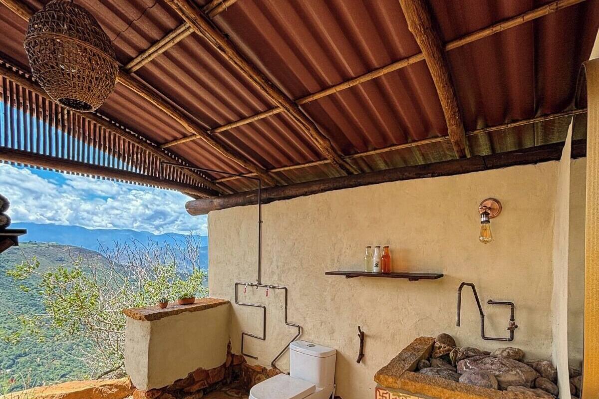 A rustic bathroom with a stone floor, a white toilet, a sink with a decorative tile backsplash, and a hanging light fixture.