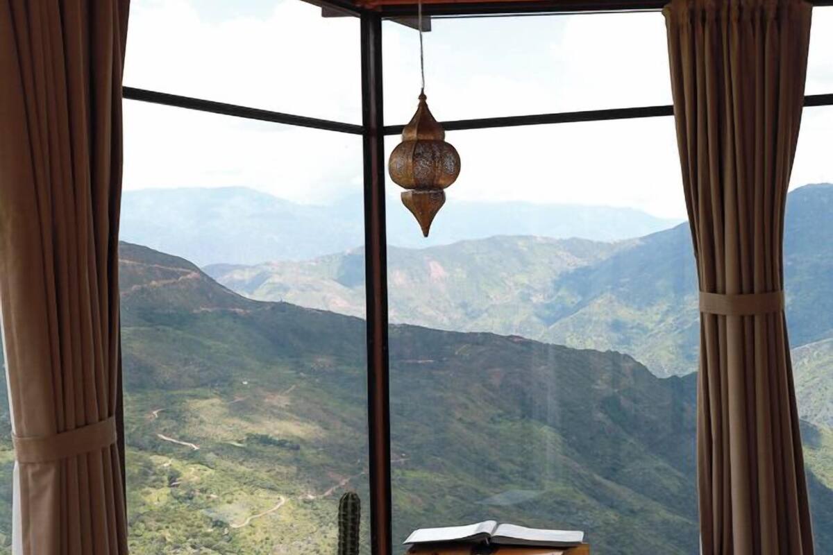A room with a wooden ceiling, a bed, a suitcase, a potted cactus, and a view of a mountainous landscape through large windows.