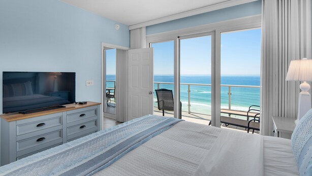 Une chambre avec un grand lit, un téléviseur posé sur une commode en bois et un balcon offrant une vue sur l’océan.