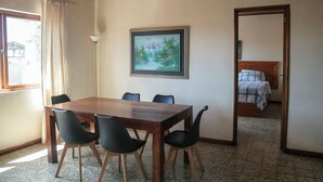 Una habitación con una mesa de comedor de madera, sillas negras, un cuadro en la pared, una cama con manta de cuadros y una ventana con cortinas.