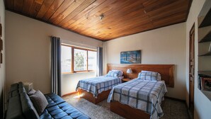 Una habitación con dos camas, un banco, una ventana con cortinas, un cuadro en la pared y un techo con vigas de madera.