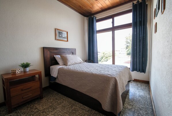 A bedroom with a wooden headboard, a bed with a beige blanket, a nightstand with a plant, and a window with curtains.