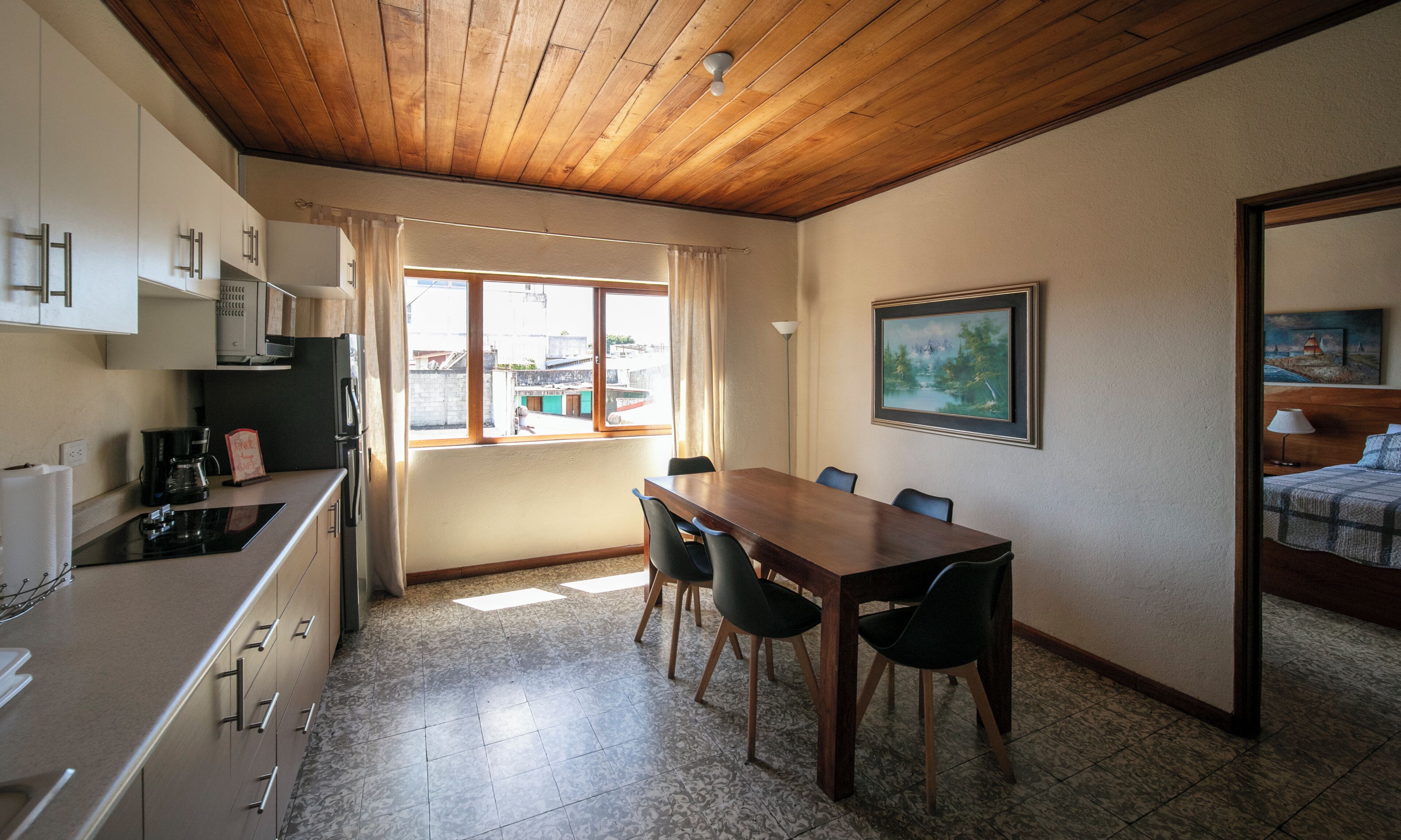 A compact kitchen with modern appliances, a dining area with a wooden table and chairs, and a large window offering a view of the outside.