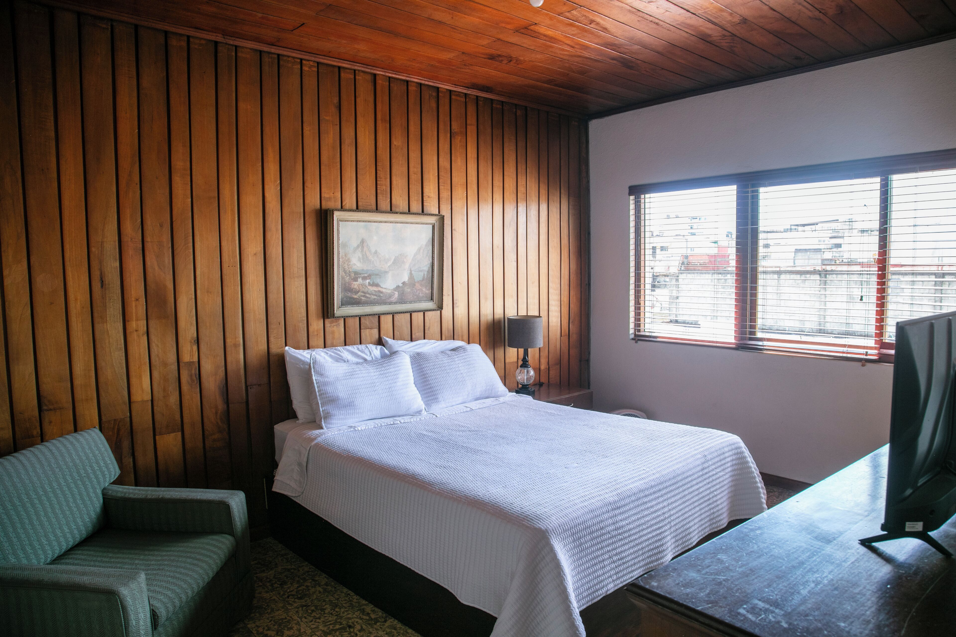 A room with a wooden paneled wall, a bed with white bedding, a green chair, a desk with a computer, and a window with blinds.