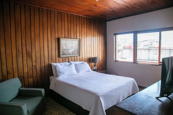 Una habitación con un revestimiento de madera, una cama con ropa blanca, una silla verde, un escritorio con una computadora y una ventana con persianas.