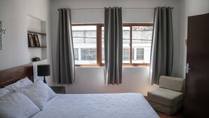 Une chambre d’hôtel avec un lit, une chaise, une table de chevet, une lampe et une fenêtre avec des rideaux.
