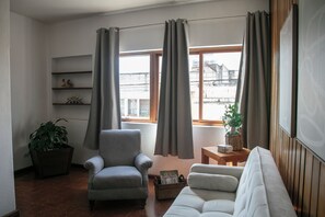 Een woonkamer met een grijze fauteuil, een witte bank, een houten salontafel, een potplant en een boekenkast met decoratieve objecten.