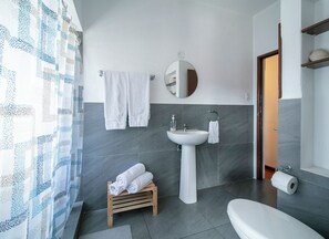 Une salle de bain moderne avec un lavabo, un miroir et des toilettes. On y trouve des serviettes sur un porte-serviettes et un petit tabouret en bois sur lequel sont également posées des serviettes.