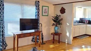 A living room with a wooden table, a television, a patterned curtain, and a kitchen in the background. - Restful manufactured suite where visitors can relax during their stay.  (Hinesville)