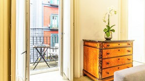 Une pièce dotée d’une porte-fenêtre coulissante qui donne sur un espace extérieur aménagé avec une table et des chaises. On y trouve une commode en bois avec des tiroirs, ainsi qu’un vase rempli de fleurs posé sur le dessus.