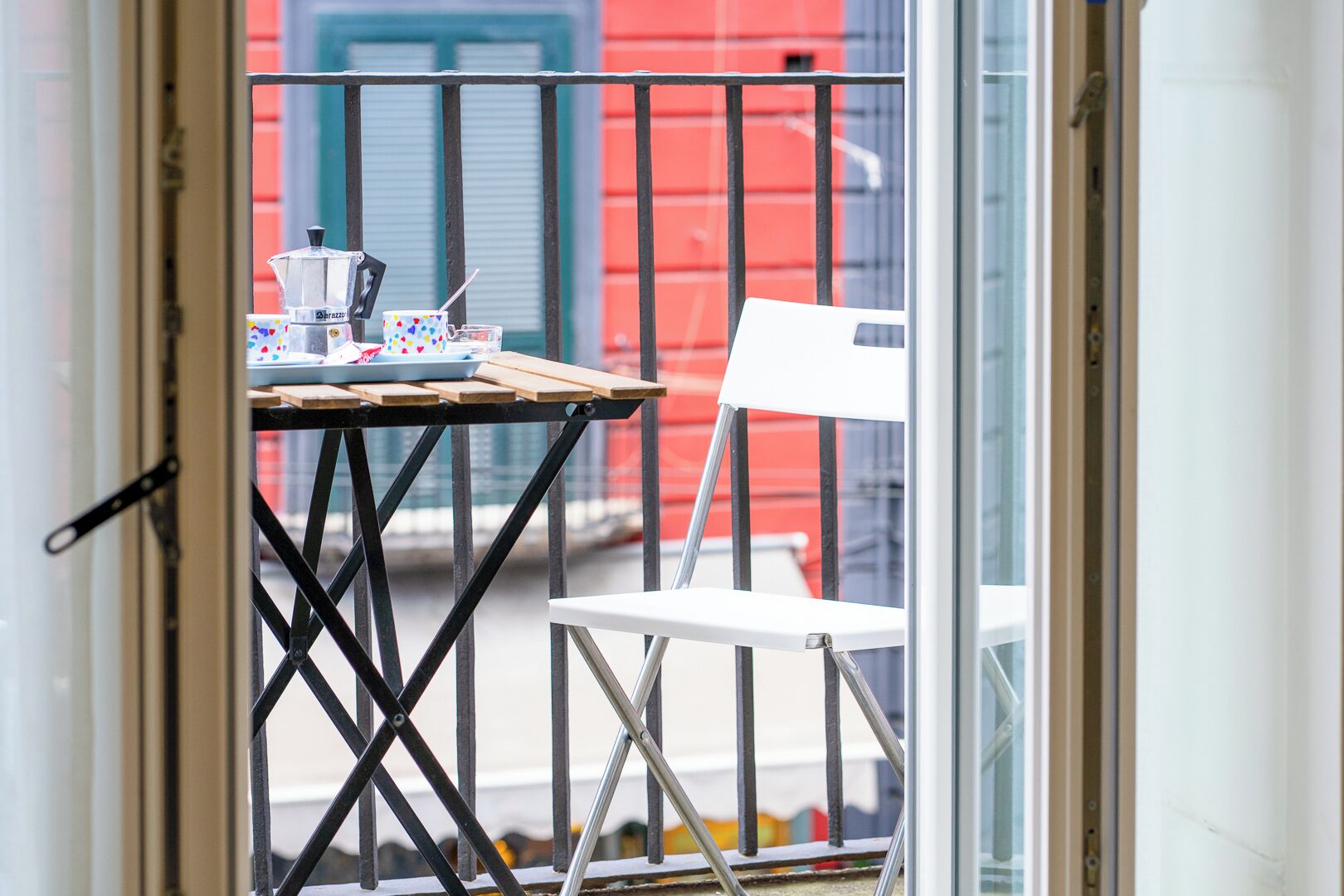 A balcony with a table and a chair, a window, and a red building in the background.