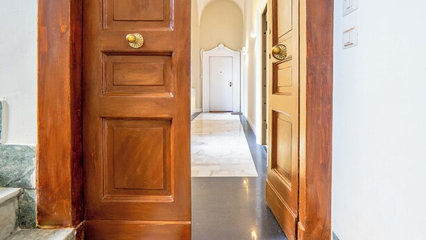 Une porte en bois dotée d’une poignée en laiton, qui donne sur un couloir au sol en marbre et aux murs blancs.
