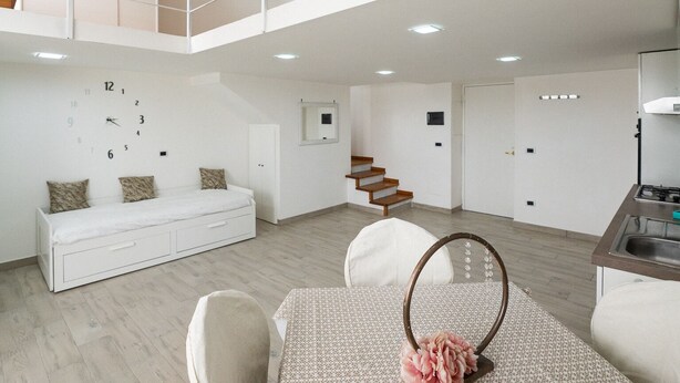 A spacious living area with a high ceiling, a staircase, a dining table with a floral centerpiece, and a white sofa with patterned cushions.