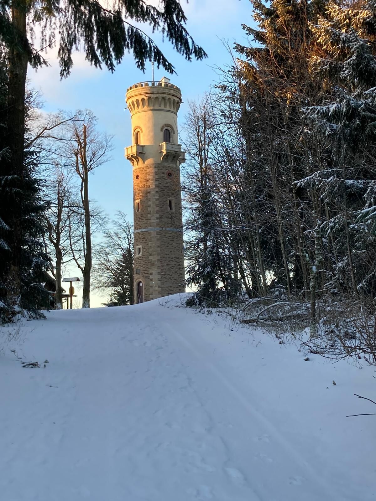 Ein verschneiter Pfad führt zu einem steinernen Turm, der von Bäumen umgeben ist.