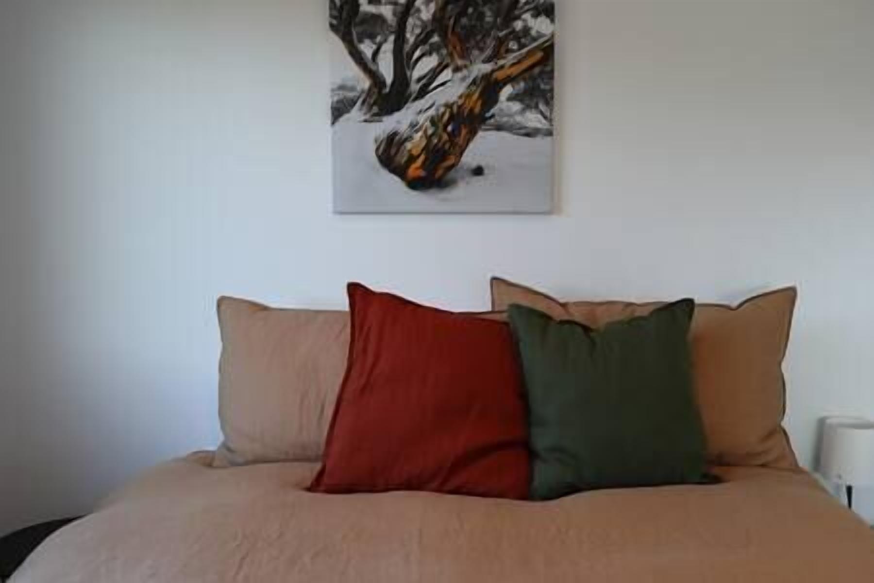 Un lit recouvert d’une couvre-lit beige et orné de deux oreillers, un rouge et un vert, dans une pièce dont le mur est blanc et au-dessus du lit, on peut voir un tableau représentant un paysage enneigé.