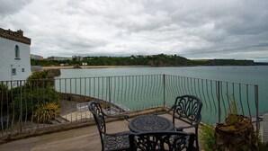 Varanda com mesa e cadeiras, vista para um corpo de água, um edifício branco e uma linha costeira distante.