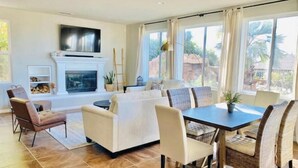 A modern living room with a fireplace, a dining area with a table and chairs, and large windows offering a view of the outdoors. - Santa Clarita Tropical Paradise, Six Flags Magic Mountain, LA, Beaches Drivable (Santa Clarita)