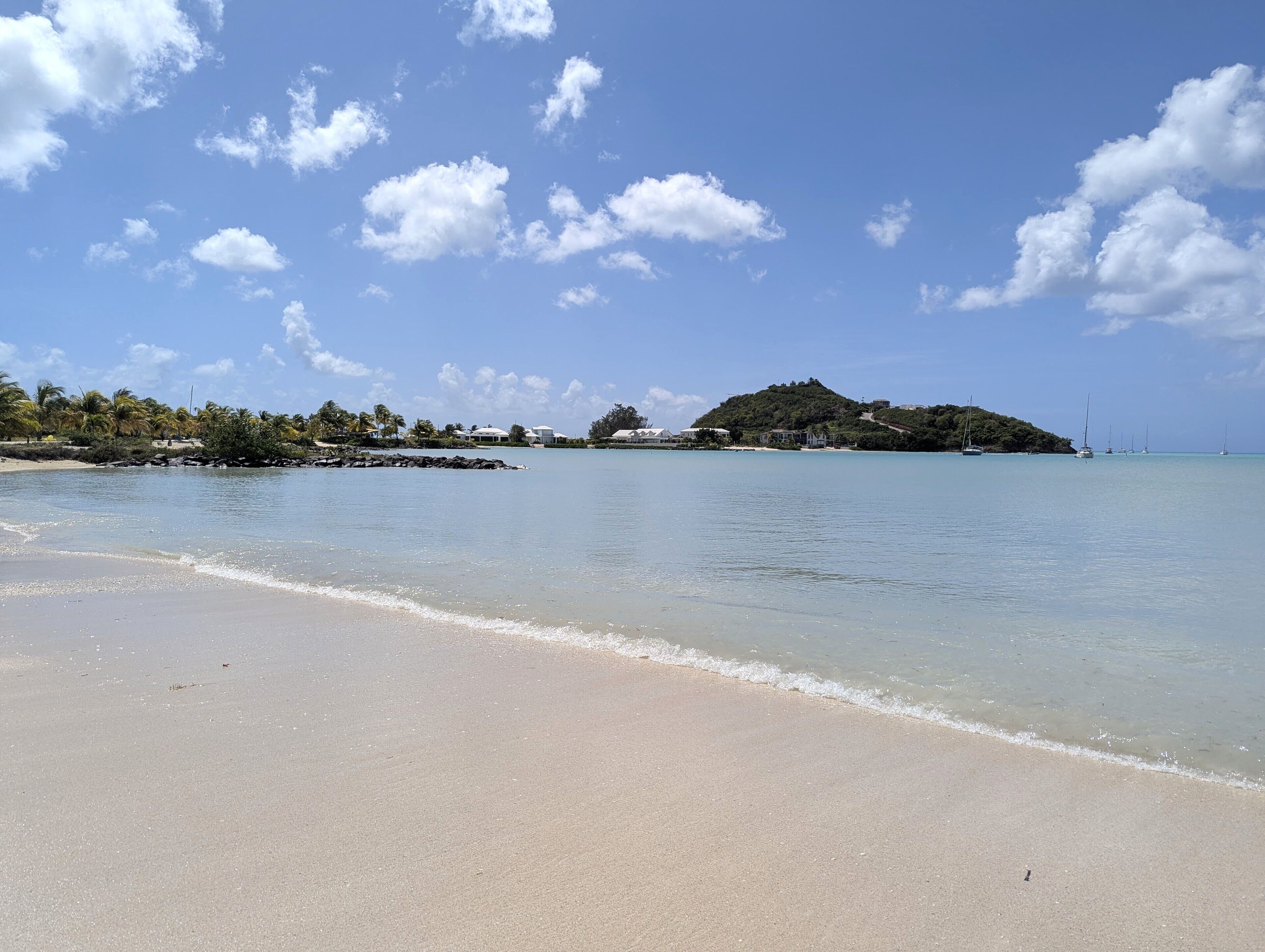 A beach with clear water, a sandy shore, and a small hill with buildings in the background.