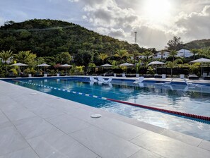 A swimming pool with clear blue water, surrounded by lounge chairs and umbrellas, set against a backdrop of lush greenery and a hill.