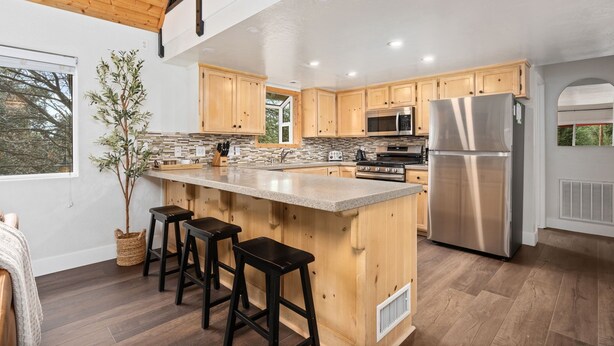 A modern kitchen with wooden cabinets, a large island with stools, stainless steel appliances, and a view of the outdoors.