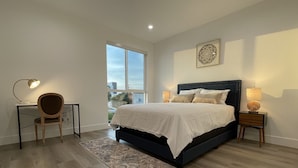 A bedroom with a large bed, a desk with a chair, a standing lamp, and a window with a view of buildings.