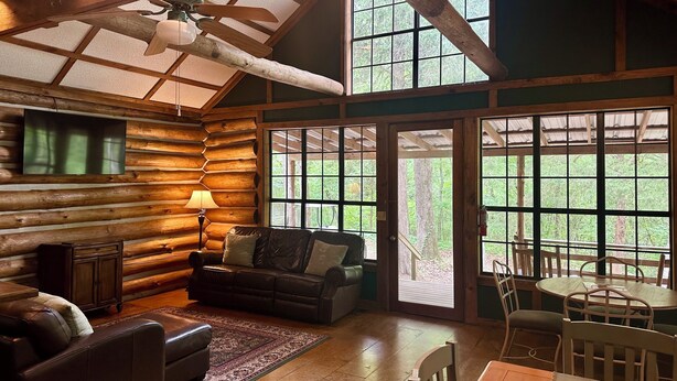 A log cabin interior with a wooden ceiling, a television mounted on the wall, a leather sofa, a dining table with chairs, and large windows offering a view of the outdoors.