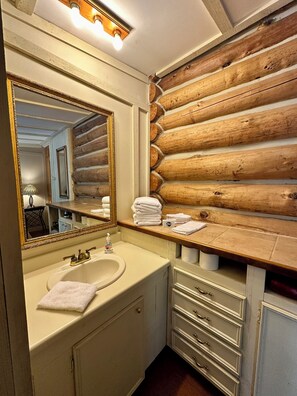A bathroom with a wooden log wall, a large mirror, a sink with a towel, and a vanity with drawers. - Antlers - An authentic log cabin experience in the forest at Stag Leap Cabins! (Nacogdoches)