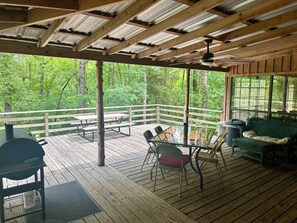A covered outdoor deck with picnic tables, chairs, and a wicker sofa, surrounded by a wooden railing and overlooking a forested area. - Antlers - An authentic log cabin experience in the forest at Stag Leap Cabins! (Nacogdoches)