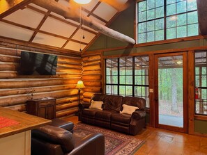 L'intérieur d'une cabane en rondins, avec un canapé en cuir, un téléviseur fixé au mur et une grande fenêtre donnant sur une forêt.