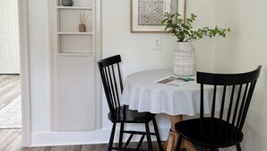A dining area with a white table, two black chairs, a white shelf with decorative items, and framed pictures on the wall.