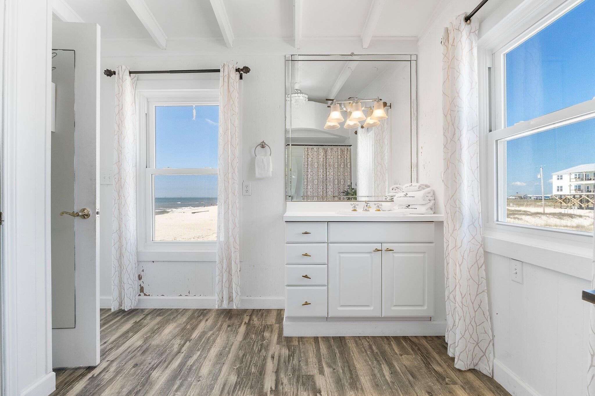 A bathroom with a wooden floor, white cabinets, a large mirror, and a view of the beach through the window.