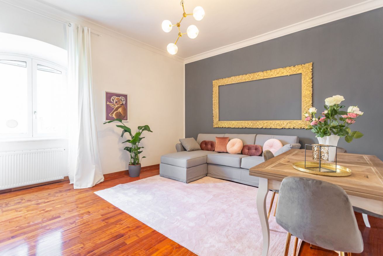 Ein modernes Wohnzimmer mit einem grauen Ecksofa, einem Holzesstisch mit goldener Zarge, einem grauen Sessel und einer Topfpflanze.