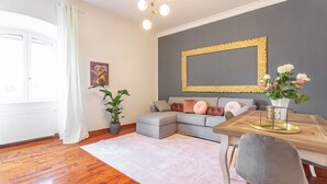A modern living room with a grey sectional sofa, a wooden dining table with a gold frame, a grey armchair, and a potted plant. - Daplace - Alma Apartment (Roma)