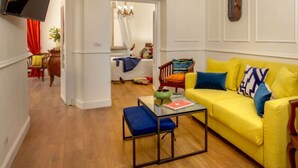 A bright yellow sofa with blue and multicolored cushions, a wooden chair, and a glass coffee table in a well-lit living room with wooden flooring and white walls. - Daplace - Flaminia Apartment (Roma)