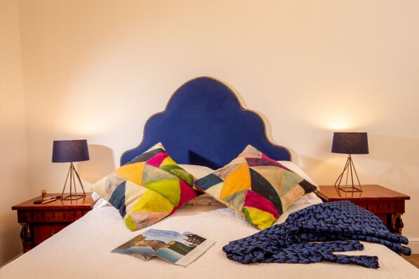 A neatly made bed with a blue headboard, colorful pillows, a blue blanket, and a magazine on the bed. There are two bedside tables with lamps and a small wooden nightstand with a lamp.