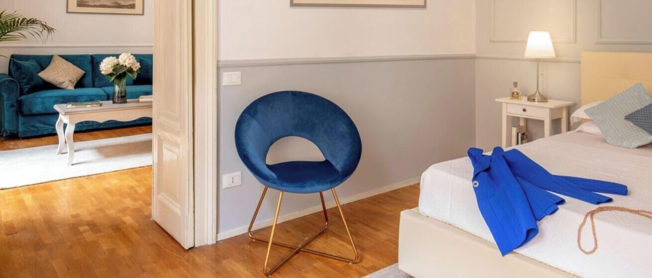 Une chambre d’hôtel avec une chaise bleue, un lit recouvert d’une couverture bleue, une table de chevet blanche avec une lampe, et un canapé bleu en arrière-plan.