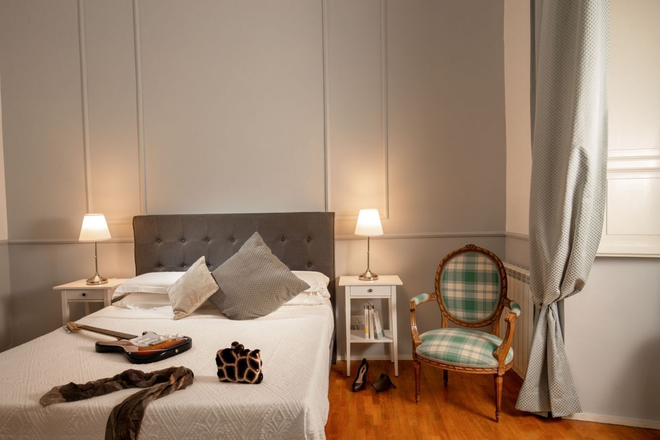 A neatly made bed with a grey headboard, two bedside lamps, a wooden chair with a plaid cushion, and a small bedside table with books.