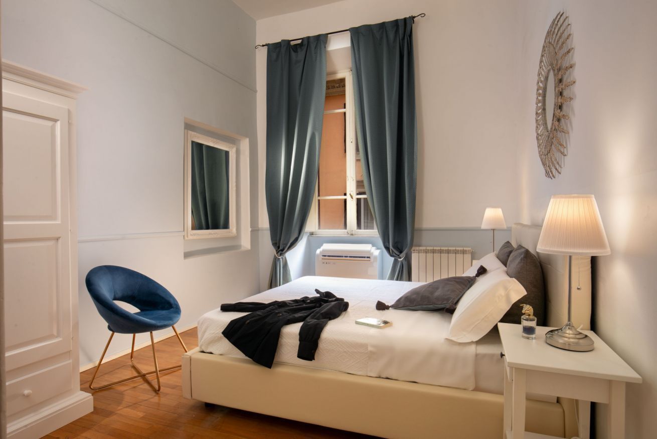 Ein Schlafzimmer mit Bett, Stuhl, Nachttisch mit Lampe, Fenster mit Vorhängen und Heizkörper.