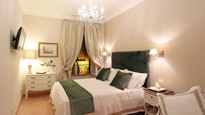 Quarto com cama grande, lustre, mesa de cabeceira com abajur, TV na parede e janela com cortinas.