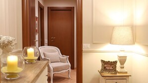 Um corredor com porta de madeira, uma poltrona branca, uma mesa com velas e uma luminária na parede.