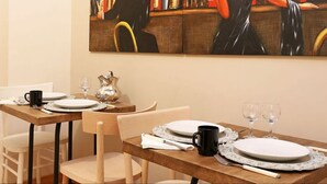 Uma área de refeições com mesas de madeira preparadas para dois, pratos brancos, canecas pretas e copos de vinho transparentes.